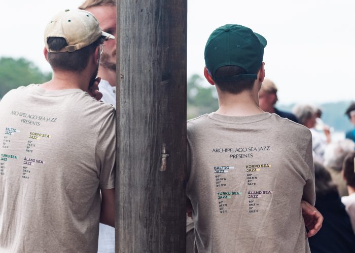 Two men with caps lean on a pilar. They are wearing grey Archipelago Sea Jazz t-shirts.